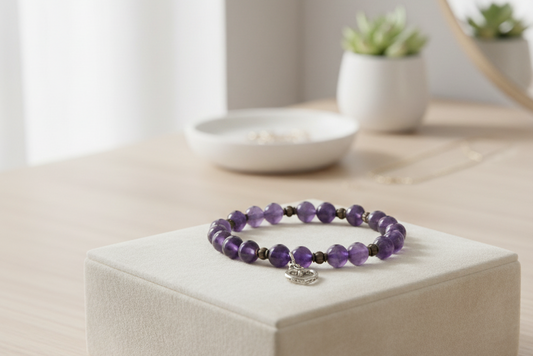Amethyst bracelet of purple beads with small dark spacers and a silver heart charm, displayed on a cream fabric block