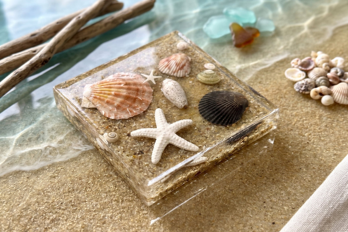 Close-up of a Coastal Treasure square resin coaster with a scallop shell, black shell and starfish, set on beach sand