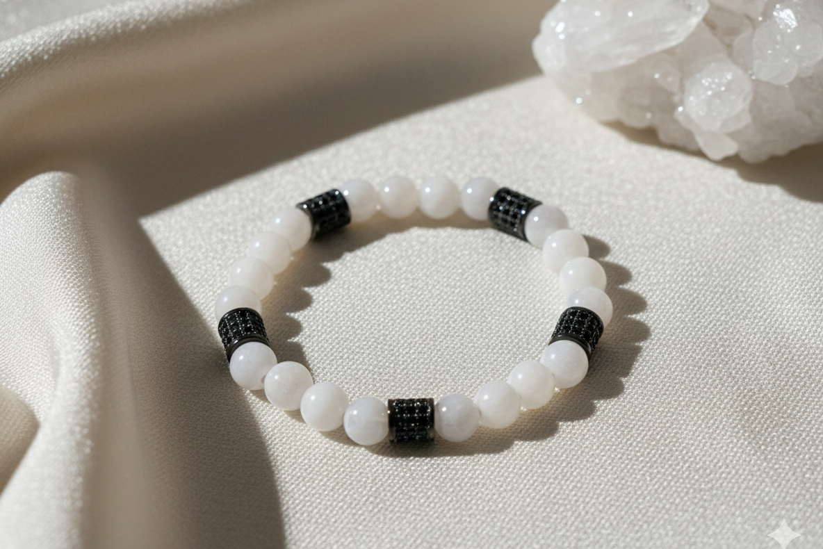 Moonstone bracelet of white beads with five black sparkly barrel spacers, laid on cream fabric in sunlight beside a clear crystal