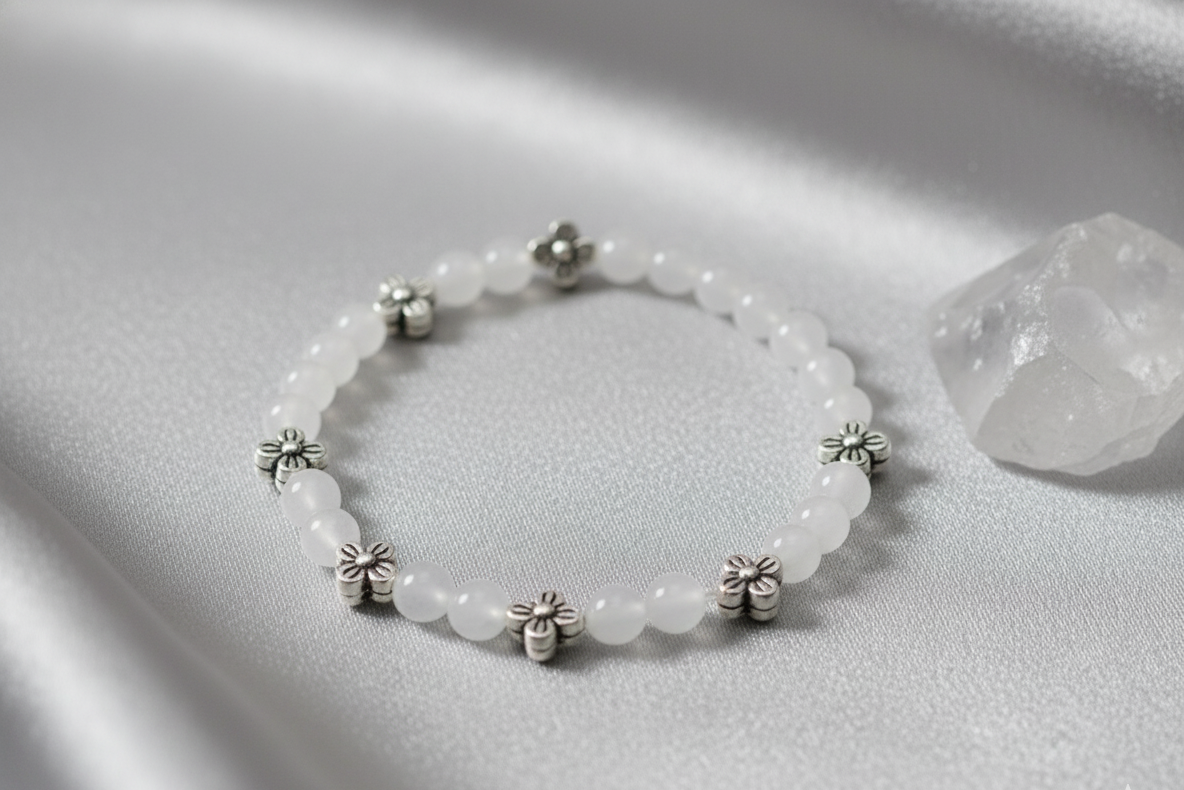 Moonstone bracelet of white beads spaced with small silver flower charms, laid on gray satin beside a clear quartz crystal